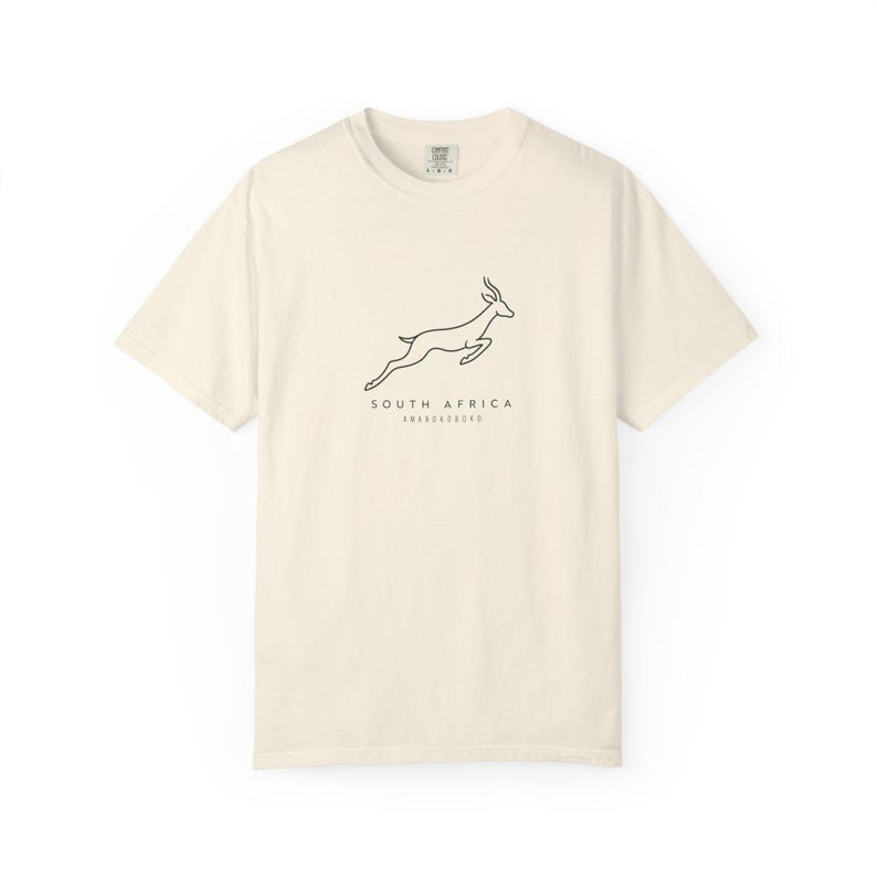 A cream-colored t-shirt with a black outline of a jumping springbok and the words "SOUTH AFRICA AMABOKOBOKO" below it, laid flat on a white background.