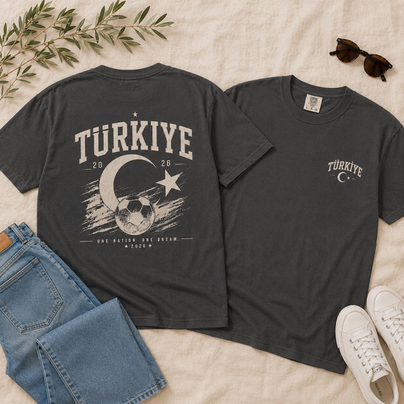 May include: A charcoal gray t-shirt featuring a large back graphic with the text "TURKIYE 20 26", a crescent moon, a star, and a soccer ball, with the text "ONE NATION ONE DREAM 2026". The front chest area displays a smaller "TURKIYE" logo with a crescent moon and star.