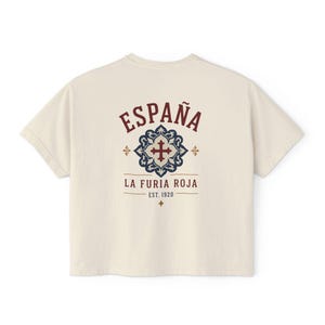 The back of a cream-colored short-sleeved crop top, showing "ESPAÑA LA FURIA ROJA EST. 1920" in red and blue with a tile graphic, shown flat against a white background.