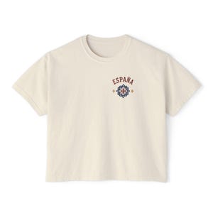 A cream-colored short-sleeved crop top with "ESPAÑA" and a red and blue tile graphic on the left chest, shown flat against a white background.