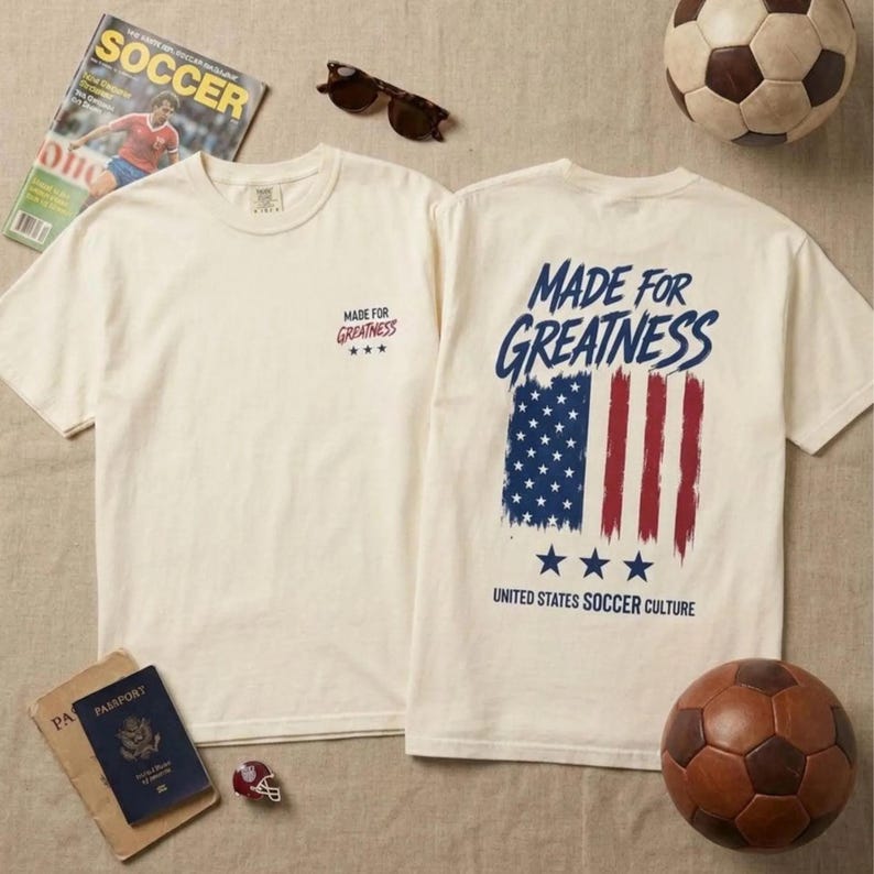 以下が含まれることがあります： オフホワイトの半袖Tシャツ。胸元には「Made for Greatness」の文字と3つの星が小さくプリントされています。背面には、かすれたアメリカ国旗の大きなグラフィックと「Made for Greatness」「United States Soccer Culture」の文字がデザインされています。