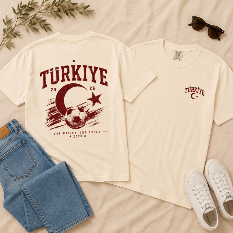 May include: An off-white t-shirt featuring a dark red graphic print. The back displays the text T&Uuml;RKIYE, 20 26, a crescent moon, a star, a soccer ball, and the text ONE NATION ONE DREAM 2026. The front chest features a small T&Uuml;RKIYE and crescent moon.
