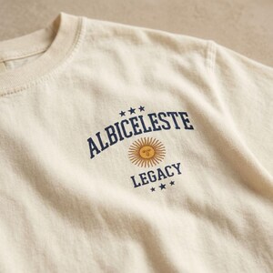 May include: A cream-colored t-shirt featuring a dark blue graphic print on the chest. The text reads ALBICELESTE LEGACY in a bold, arched font, accompanied by a central yellow sun icon and three small stars above and below the text.