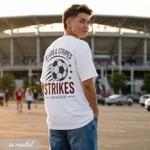 May include: A white short-sleeve t-shirt featuring a graphic on the back with a soccer ball, red and blue motion lines, and the text "STARS & STRIPES STRIKES USA 2026". The design includes stars and horizontal lines in a patriotic color scheme.