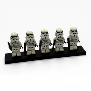 May include: Five white and black plastic stormtrooper minifigures standing in a row on a rectangular black display base. Each figure wears a detailed helmet and armor with black accents and holds a small black blaster accessory in one hand.