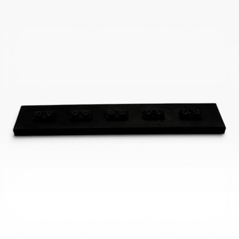 May include: A rectangular black plastic building block base plate featuring five distinct two-stud connection points arranged in a single horizontal row across the surface. The object is solid black with a matte finish and a flat, elongated design.
