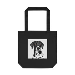May include: Black tote bag featuring a square image of a dog with black and white spots. The dog has a black head and ears, with brown eyes. The tote bag has two black handles.