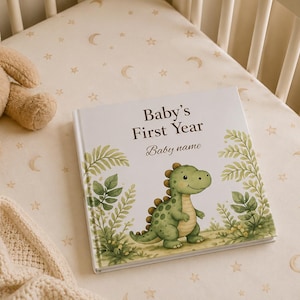 May include: A square hardcover book titled Baby's First Year with a placeholder for a baby name. The cover features a green cartoon dinosaur standing among green ferns and foliage. The book is placed on a cream-colored surface with stars.