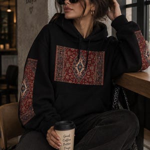May include: A black hooded sweatshirt featuring a rectangular patch on the chest and matching panels on the sleeves, all displaying a detailed red, blue, and cream-colored ornate rug pattern. The hoodie includes a front kangaroo pocket and drawstrings.