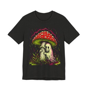 Può includere: T-shirt nero con una grafica colorata di un fantasma che si ripara sotto un fungo rosso e bianco sotto la pioggia. Il fungo ha un lato inferiore verde e il fantasma è bianco con una sfumatura rosata. Il design è su erba verde e rosa.