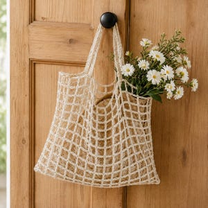 May include: A cream-colored crocheted net tote bag with an open square grid pattern and two integrated handles. The bag contains a bouquet of white daisies with yellow centers and green stems, hanging from a dark knob on a wooden door panel.