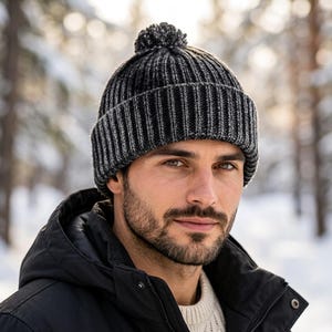 Könnte beinhalten: Eine anthrazitfarbene und schwarze, gerippte Strickmütze mit einem passenden Bommel an der Oberseite. Die Mütze verfügt über ein dickes, umgeschlagenes Bündchen und ein strukturiertes, vertikales Strickmuster für einen klassischen Look.