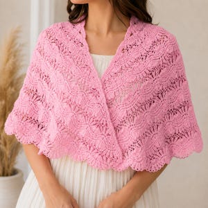 May include: A light pink crochet shawl featuring an intricate openwork pattern with scalloped edges. The garment is draped over the shoulders, showcasing a delicate, textured knit design that covers the upper torso and arms with a soft, airy appearance.