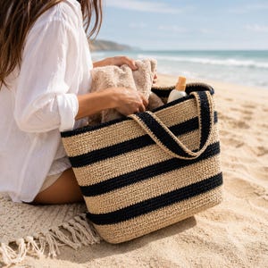 May include: A large, rectangular tote bag featuring horizontal stripes in natural beige and dark navy blue. The bag is constructed from a woven material and includes two matching striped shoulder straps, containing a beige towel and a bottle.