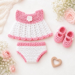 May include: A handmade crochet baby outfit in white and pink. The set includes a dress, diaper cover, and a pair of Mary Jane shoes. A wooden heart and flowers are also in the image.