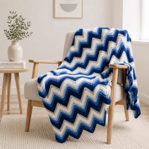 Puede incluir: Una manta de ganchillo con un patrón de chevrón en tonos azul marino, azul real, azul claro y crema. El tejido está drapeado sobre un sillón de color claro, mostrando el diseño de punto en zigzag y la transición gradual de los colores.