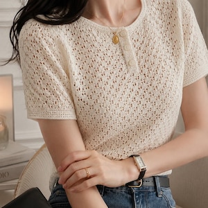 May include: A cream-colored short-sleeved top featuring an intricate openwork knit pattern, a round neckline, and a vertical placket with three matching buttons. The garment is paired with blue denim jeans and a black leather belt with a gold buckle.