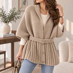 May include: A beige knit cardigan featuring a shawl collar, three-quarter length sleeves, and a matching knit belt secured with four round, textured buttons down the front. The garment is worn over a white shirt and light blue denim jeans.