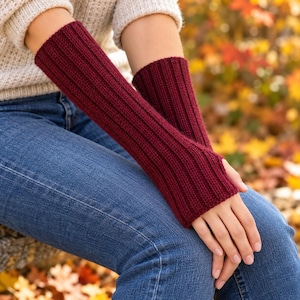 May include: A pair of deep burgundy fingerless gloves featuring a vertical ribbed knit pattern. The gloves extend from the knuckles to the mid-forearm, crafted with a consistent texture and a snug fit around the hands and wrists.