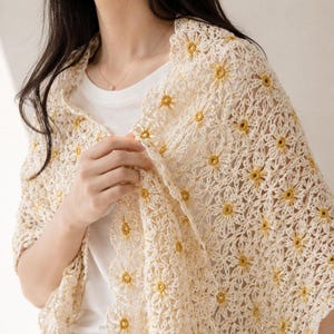 Peut inclure: Un châle en crochet de couleur crème présentant un motif floral répétitif avec des centres jaunes. Le design délicat et ajouré est composé de motifs en forme de marguerites interconnectés, créant un bord festonné le long du vêtement.
