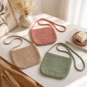 May include: Three crochet crossbody bags in shades of beige, dusty rose, and sage green are displayed on a white surface. Each bag has a matching strap. The bags are rectangular with a textured, woven appearance, suggesting a handmade craft.