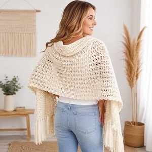 May include: A cream-colored crochet shawl featuring an openwork grid pattern, a scalloped edge, and long fringe detailing along the bottom hem. The garment is draped over the shoulders and hangs down the back, paired with light blue denim jeans.