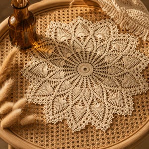 May include: A round, cream-colored crochet doily featuring an intricate, radiating floral pattern with a central circular motif. The lace-like textile displays detailed stitching and scalloped edges, creating a delicate, symmetrical design.