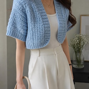 Puede incluir: Un cárdigan de punto azul claro de manga corta con patrón texturizado y bordes acanalados, usado sobre una camiseta sin mangas blanca lisa y combinado con pantalones blancos de talle alto. El cárdigan tiene un diseño abierto con cuello ancho.