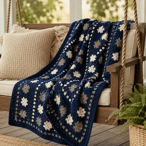 Op de afbeelding: Een handgehaakte deken met een herhalend granny square patroon in diep marineblauw, crème en zachtgrijs garen. Het textiel toont een geometrische rangschikking van kleine bloemachtige motieven, afgewerkt met een textuurrijke rand.