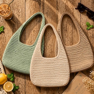 Könnte beinhalten: Drei gehäkelte Hobo-Taschen in Salbeigrün, Creme und Hellbraun. Jede Tasche verfügt über einen einzelnen integrierten Schulterriemen und ein strukturiertes, horizontal geripptes Strickmuster. Die Taschen sind gruppiert angeordnet.