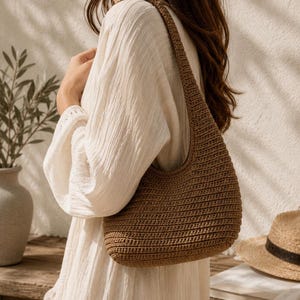Peut inclure: Un sac &agrave; bandouli&egrave;re marron de style hobo, r&eacute;alis&eacute; avec un motif en maille crochet&eacute;e dense et textur&eacute;e. Le sac pr&eacute;sente une sangle unique int&eacute;gr&eacute;e et une silhouette arrondie, mettant en valeur un point uniforme sur tout l'ext&eacute;rieur.