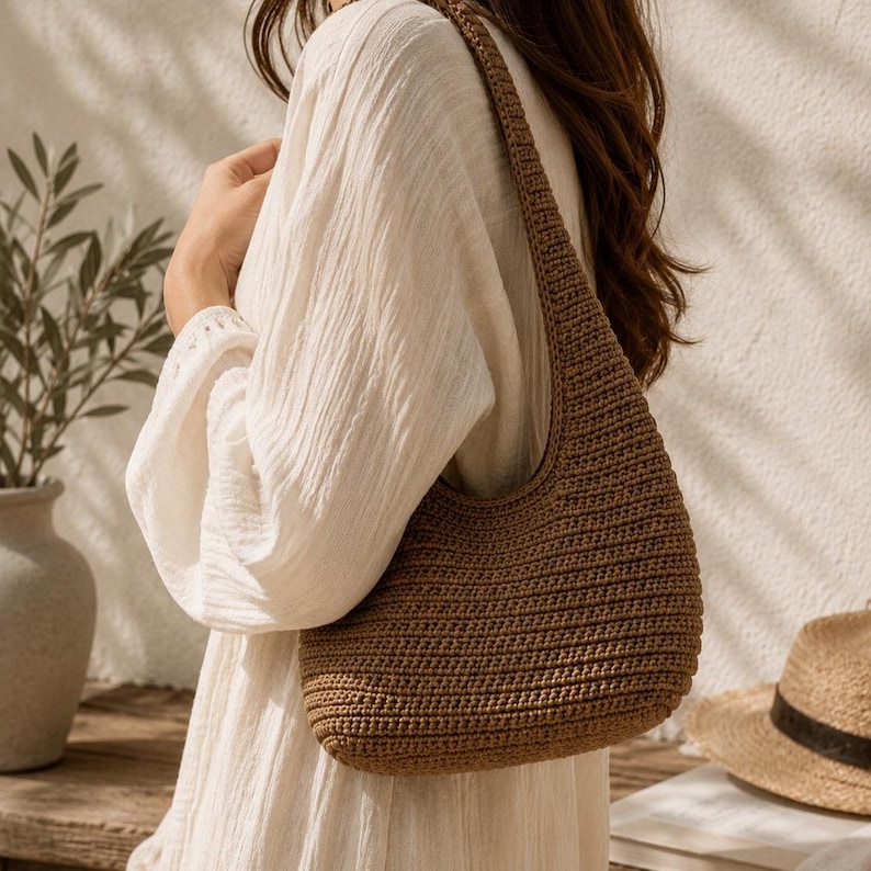 Peut inclure: Un sac &agrave; bandouli&egrave;re marron de style hobo, r&eacute;alis&eacute; avec un motif en maille crochet&eacute;e dense et textur&eacute;e. Le sac pr&eacute;sente une sangle unique int&eacute;gr&eacute;e et une silhouette arrondie, mettant en valeur un point uniforme sur tout l'ext&eacute;rieur.