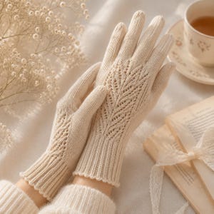 May include: A pair of cream-colored knit gloves featuring a delicate, openwork leaf pattern on the back of the hand and a ribbed cuff. The gloves are crafted with a fine-gauge knit texture, showcasing a subtle, intricate design that extends from the wrist.