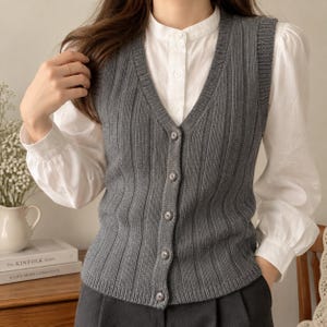 May include: A dark grey sleeveless knit vest featuring a vertical ribbed texture, a V-neckline, and a front button closure with five matching circular buttons. The vest is layered over a white long-sleeved shirt with a mandarin collar and cuffs.