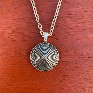 Könnte beinhalten: Eine silberne Halskette mit einem runden Anhänger, der ein konzentrisches Kreisdesign aufweist. Der Anhänger hat eine strukturierte Oberfläche und eine silberne Kette.