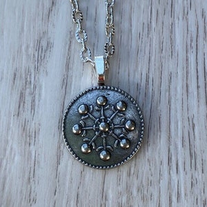 Puede incluir: Un collar de cadena de plata con un colgante redondo que presenta un diseño de copo de nieve estilizado. El colgante tiene un borde de cuentas y una superficie texturizada.