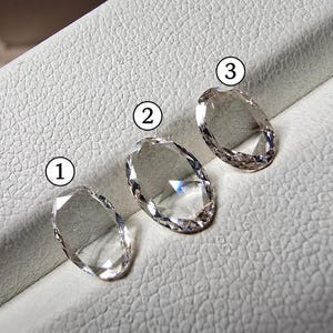 Puede incluir: Tres piedras preciosas transparentes de corte ovalado dispuestas en fila, cada una etiquetada con una pequeña etiqueta circular blanca con un número negro. Las piedras están numeradas 1, 2 y 3, mostrando superficies facetadas que reflejan la luz.