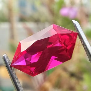 May include: A vibrant, deep pink, faceted gemstone with a hexagonal, elongated shape. The stone features sharp, geometric cuts that create multiple light-reflecting surfaces, held securely by a pair of metal tweezers against a blurred outdoor background.