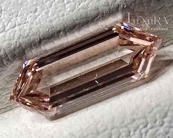 May include: A loose, elongated hexagonal gemstone with a light peach or champagne hue. The stone features a step-cut facet pattern that creates sharp, geometric lines and internal reflections. The gem is positioned on a textured white surface.