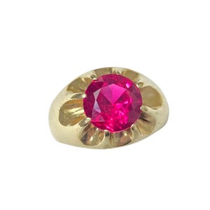 Puede incluir: Un anillo de oro con una gran gema roja brillante y redonda. El anillo tiene un diseño clásico con una banda lisa y pulida y un engaste decorativo alrededor de la gema. La gema es el punto focal, captando la luz y añadiendo un toque de elegancia.