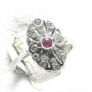 May include: A silver ring with an oval-shaped face. The face features a central ruby surrounded by numerous small diamonds. The ring's design is intricate, with the diamonds set in a radial pattern around the ruby.