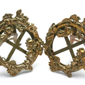 Könnte beinhalten: Zwei verzierte goldfarbene Bilderrahmen aus Metall mit einem kreuzförmigen Design in der Mitte. Die Rahmen sind mit einem floralen Motiv verziert.