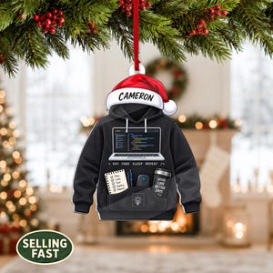 May include: A black hoodie ornament featuring a red Santa hat with the name CAMERON, a laptop graphic with code, a notepad with a checklist, a pen, a computer mouse, and a tumbler with the text EAT CODE SLEEP REPEAT and GOOD CODE BETTER DAYS.