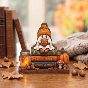 Può includere: Una statuetta decorativa raffigurante un fantasma bianco con un berretto di lana arancione che tiene una tazza con la scritta "SWEATER WEATHER", seduto su una pila di tre libri intitolati "COZY NIGHTS", "FALL WHISPERS" e "WOODLAND MAGIC".