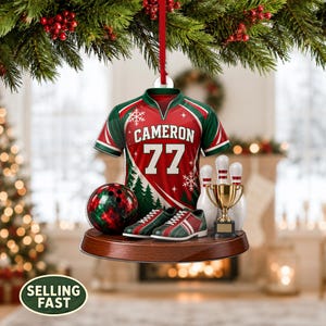 Op de afbeelding: Een ornament hangend aan een rood lint aan een dennentak, met een rood en groen bowling-shirt met de naam CAMERON en nummer 77, een bowlingbal, een paar bowlingschoenen en een gouden trofee op een houten voetstuk.
