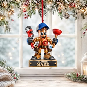 May include: A hanging Christmas ornament featuring a cartoon plumber character made of copper pipes, wearing a blue cap and brown work boots. The figure holds a red pipe wrench and a red plunger, standing on a base labeled PLUMBER MARK.