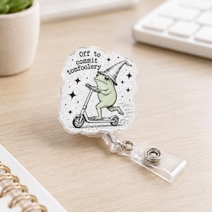 May include: A clear acrylic badge reel featuring a glittery finish and a printed illustration of a light green frog wearing a pointed hat while riding a scooter. The text "Off to commit tomfoolery" is printed above the frog with small star accents.