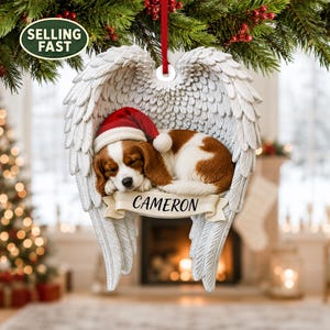 Könnte beinhalten: Ein Weihnachtsschmuck mit einem schlafenden braun-weißen Hund mit roter Weihnachtsmütze, eingebettet zwischen zwei großen weißen Engelsflügeln. Ein cremefarbenes Banner auf der Vorderseite zeigt den Namen CAMERON in schwarzen Großbuchstaben.
