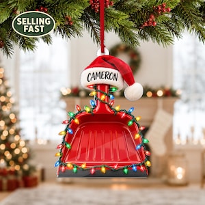 Könnte beinhalten: Ein roter Kehrblech-Weihnachtsschmuck, der an einem roten Band hängt. Das Kehrblech ist mit einer bunten Lichterkette umwickelt und mit einer rot-weißen Weihnachtsmannmütze versehen, auf deren weißem Rand der Name CAMERON in Schwarz gedruckt ist.