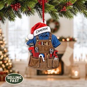 May include: A Christmas tree ornament shaped like a blue shirt and brown work apron with various tools including a pipe wrench, pliers, and tape. A red Santa hat sits on top with the name CAMERON printed in black text on the white brim.